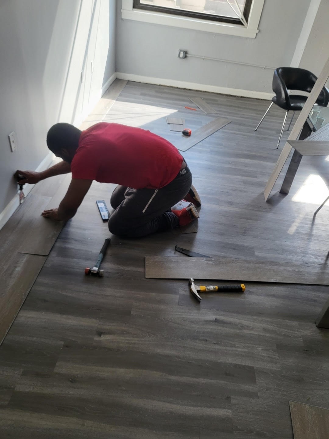 A professional installer fitting elegant wood-look vinyl plank flooring in a spacious living room. The composition showcases a bright, airy space with soft diffused lighting filtering through large windows. The color palette includes warm browns and creamy whites that create an inviting atmosphere. The camera captures the scene from a low angle, highlighting the smooth texture of the planks and the installer’s focused expression. Environmental elements include a completed section of pristine vinyl flooring, tools neatly arranged nearby, and a few potted plants adding a pop of green. The style references contemporary home decor photography, aiming for hyperrealistic detail in 8K resolution to accentuate the wood grain and shine of the vinyl surface.